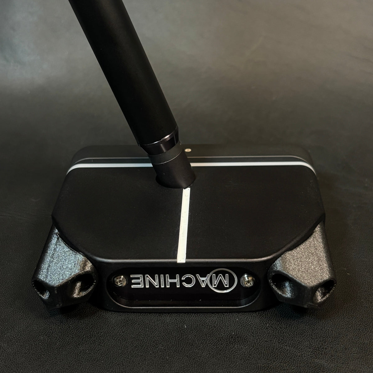 A close-up of a black golf putter head with a silver and black handle, resting on a dark textured surface. The putter features the brand name 'MACHINE' in silver lettering on the front.