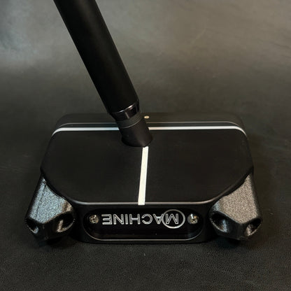 A close-up of a black golf putter head with a silver and black handle, resting on a dark textured surface. The putter features the brand name 'MACHINE' in silver lettering on the front.