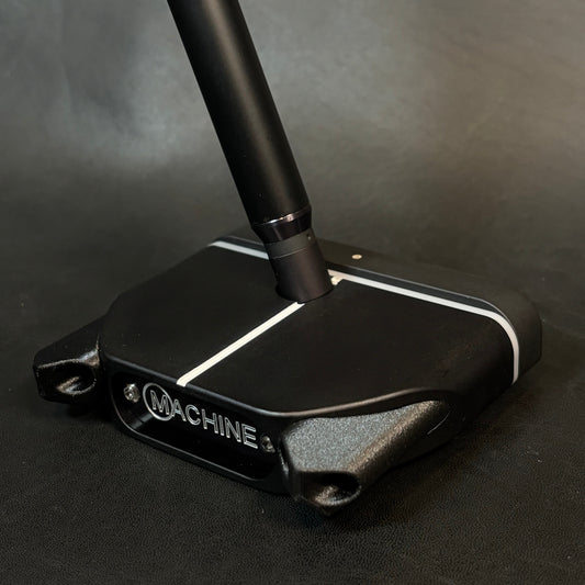 A close-up of a black golf putter head with a matching black shaft, featuring white accent lines and the visible brand name 'MACHINE' on the side, set against a dark textured background.