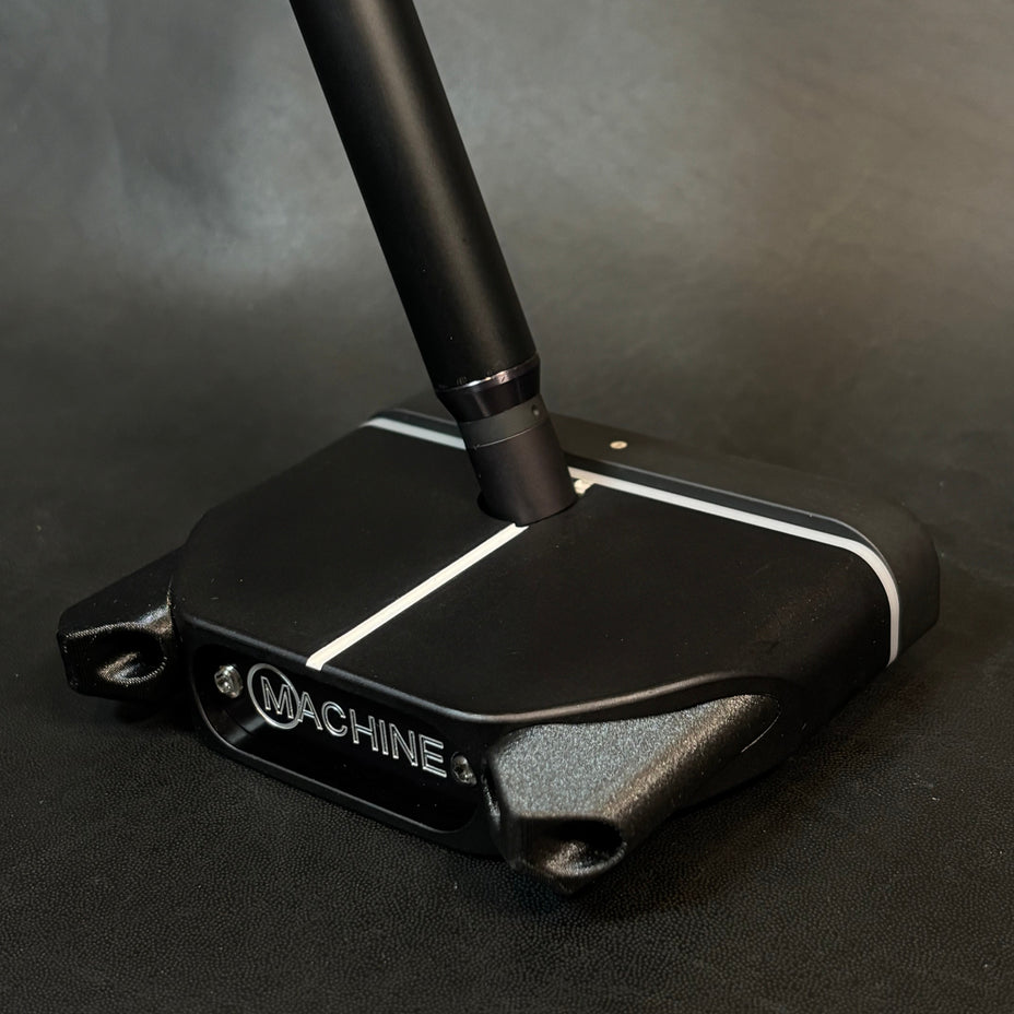 A close-up of a black golf putter head with a matching black shaft, featuring white accent lines and the visible brand name 'MACHINE' on the side, set against a dark textured background.