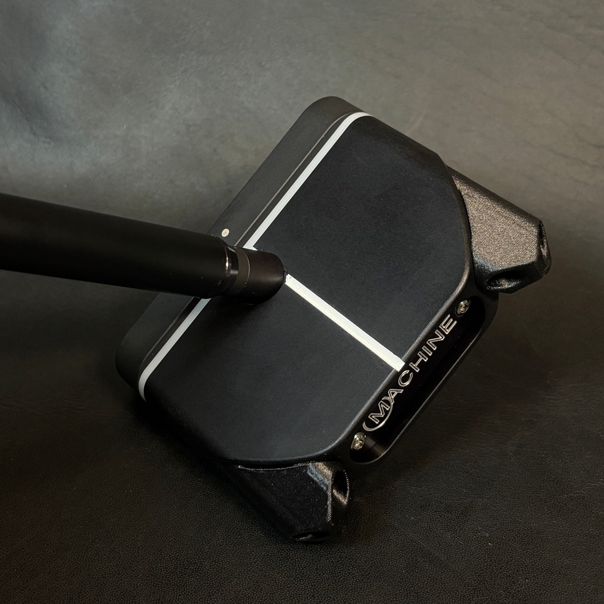 A close-up of a black golf putter head with a white center line and a black shaft, resting on a dark textured surface. The brand name 'WACHINE' is visible on the side of the club head.