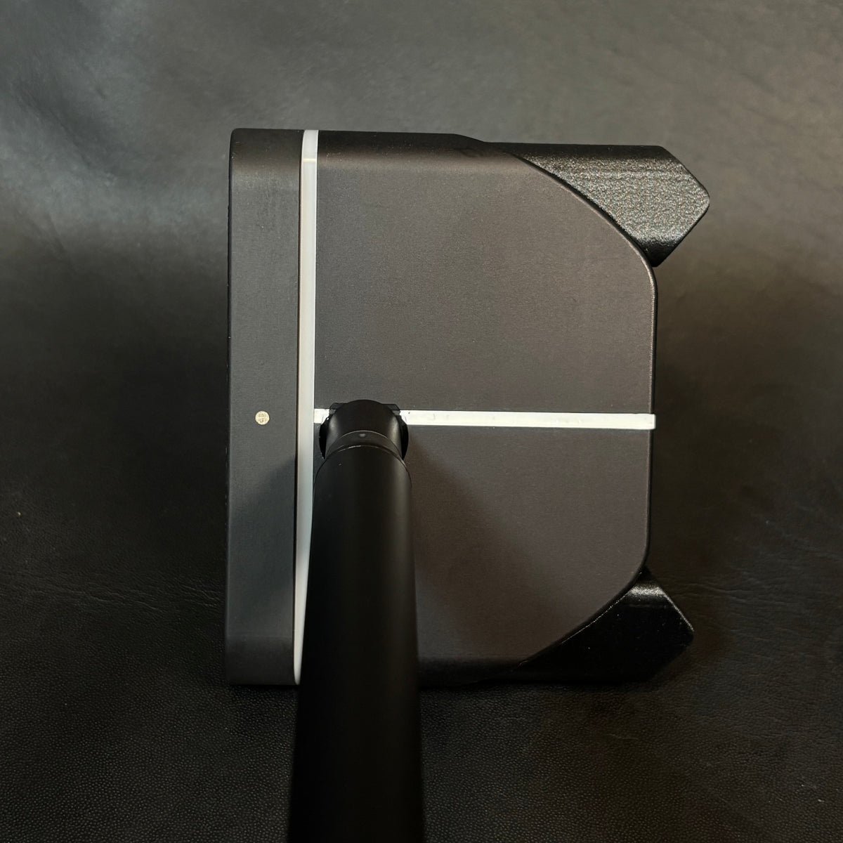 Close-up of a black golf club head and shaft, showing a matte finish with a thin white stripe detail, photographed on a dark textured surface.