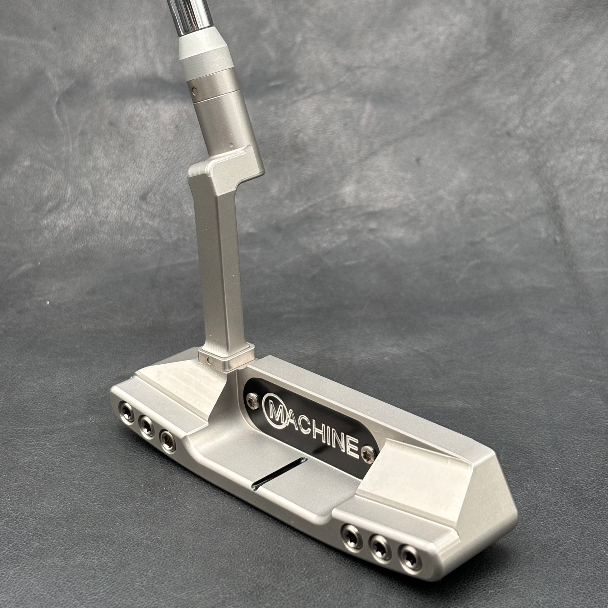 A silver metallic golf putter with a black and white 'MACHINE Q' logo plate, resting on a textured gray background.