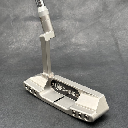 A silver metallic golf putter with a black and white 'MACHINE Q' logo plate, resting on a textured gray background.